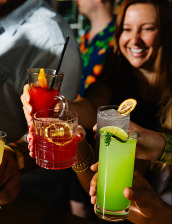 Lively bar scene with friends toasting colorful cocktails — neon-green lime drink with lime wedge, ruby-red and frozen red cocktails garnished with citrus, smiling crowd in the background.