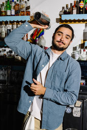 Bartender shaking a metal cocktail shaker behind a bar with shelves of liquor bottles and a small rainbow pride flag in the background.