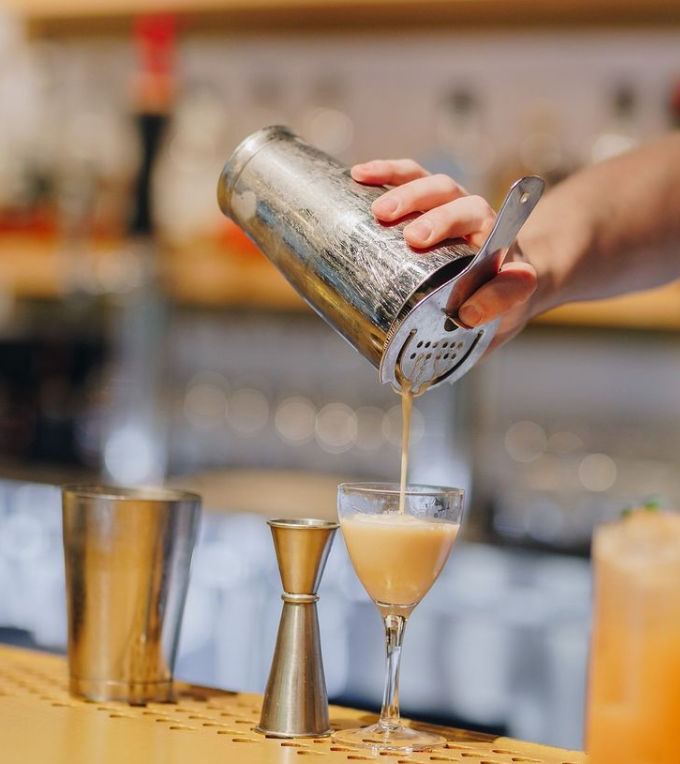Bartender pouring a frothy golden shaken cocktail through a Hawthorne strainer from a metal shaker into a coupe glass on a bar counter, with a jigger and mixing tin nearby.