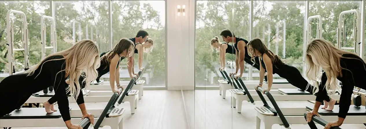 Group Pilates reformer class in a bright fitness studio, participants doing plank-style exercises on reformer machines facing a mirrored wall and large windows with a green outdoor view.