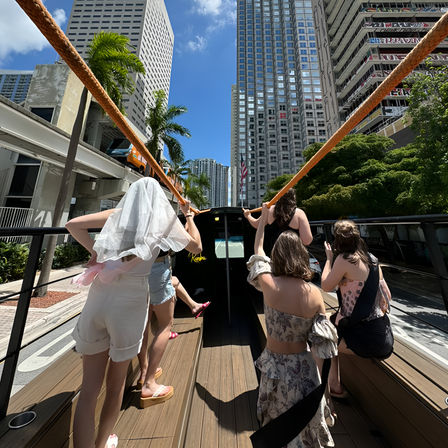 Bachelorette crew on an open-top party bus cruising downtown Miami, bride in veil holding orange strap with glass skyscrapers and palm trees overhead