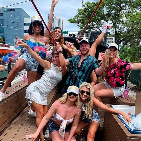 Energetic group of friends partying on an open-air trolley in Miami, wearing summer outfits, hats and sunglasses, cheering with drinks against an urban waterfront backdrop