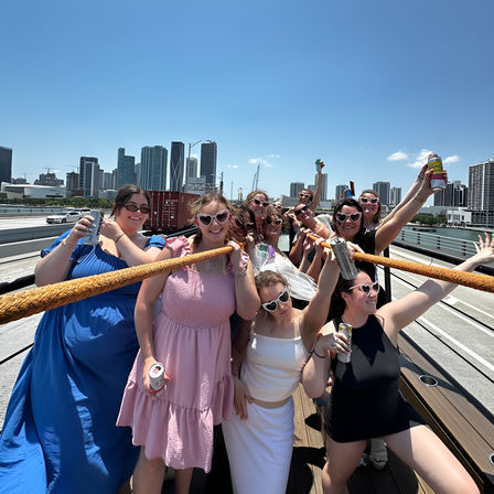 Joyful group of women on a sunny boat deck holding drinks and a thick rope, wearing summer dresses and heart-shaped sunglasses, with a waterfront city skyline and clear blue sky behind them