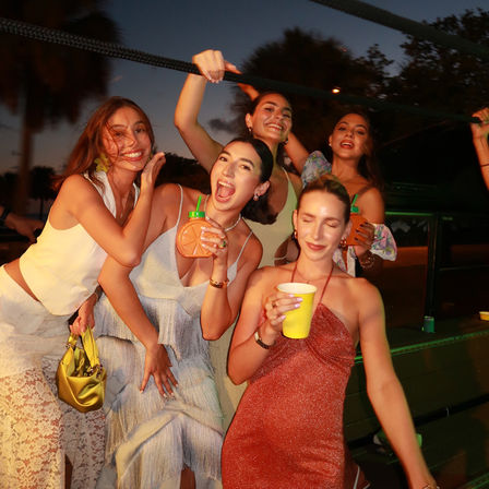 Five friends in summer dresses laughing and posing on an outdoor tropical deck at dusk with palm trees, holding colorful cups and a novelty orange-shaped drink — lively summer party vibe