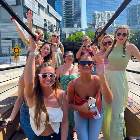 Group of smiling friends in sunglasses on an open-top party bus in a sunny downtown with glass skyscrapers, waving and holding drinks.
