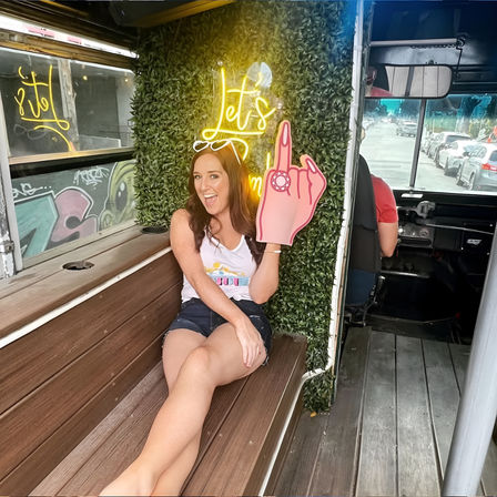 Smiling woman lounging on a wooden bench inside a party bus, holding a pink foam middle-finger prop in front of a boxwood wall with a neon “Let’s” sign, city street visible through the front window.