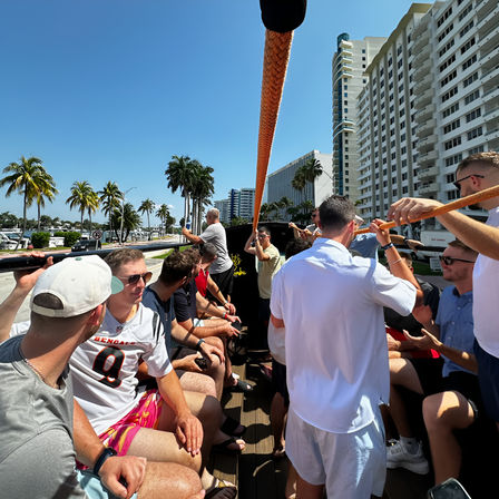 Group of friends on a sunny open-top party boat along a Florida waterfront with palm trees and high-rise condos, passengers holding an orange rope