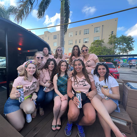 Smiling group of women in matching pink shirts and heart-shaped sunglasses enjoying a sunny Miami rooftop patio with palm trees.