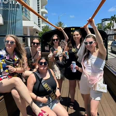 Cheerful bachelorette party of six women in sunglasses and sashes riding a wooden-deck party trolley on a sunny coastal street with palm trees and modern buildings, holding canned drinks.