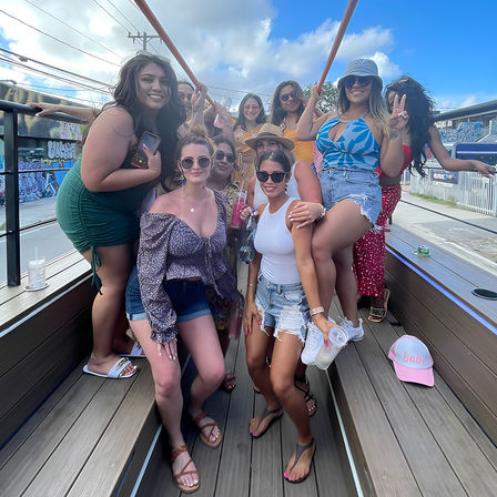 Group of smiling women on an open-air party trolley posing with drinks and sunglasses against colorful graffiti and blue sky — sunny summer girls' day out.