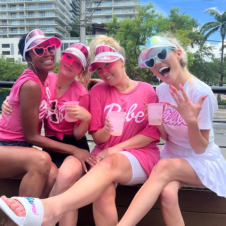 Four women in bright pink bachelorette shirts and visors, sipping drinks and laughing on a waterfront bench with palm trees and high-rise condos in the background.