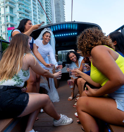 Cheerful bachelorette party on a daytime urban waterfront boat, bride in a veil and friends laughing and toasting with plastic cups against a backdrop of high-rise buildings.