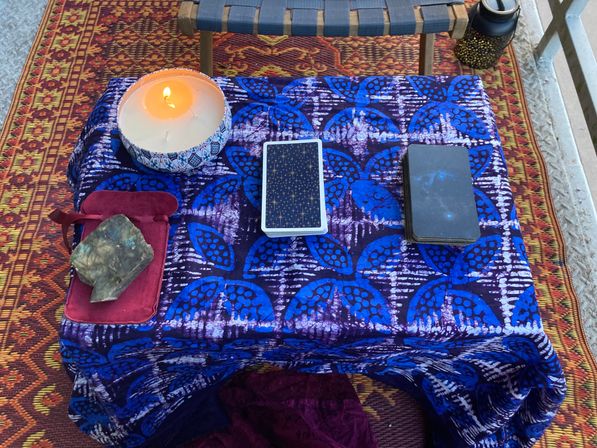 Bohemian balcony altar on patterned rug: small table draped in blue batik cloth with two tarot decks, a lit decorative candle, and a labradorite crystal on a red pouch.