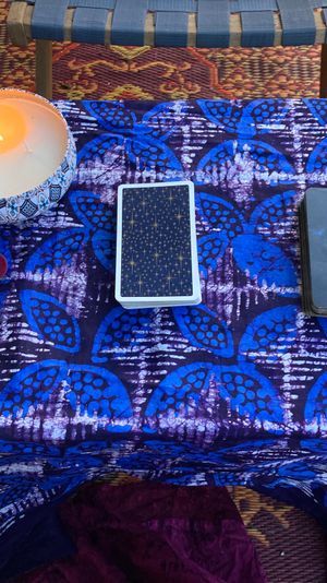 Bohemian balcony altar on patterned rug: small table draped in blue batik cloth with two tarot decks, a lit decorative candle, and a labradorite crystal on a red pouch.