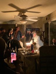 Cozy indoor birthday party at home — friends gathered around a dining table as a person leans toward a lit cake with many candles, guests cheer and snap photos with smartphones beneath a ceiling fan.