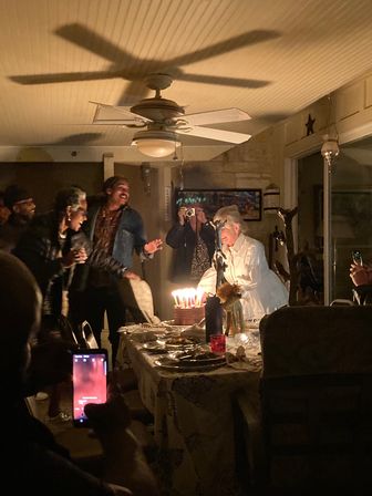 Cozy indoor birthday party at home — friends gathered around a dining table as a person leans toward a lit cake with many candles, guests cheer and snap photos with smartphones beneath a ceiling fan.