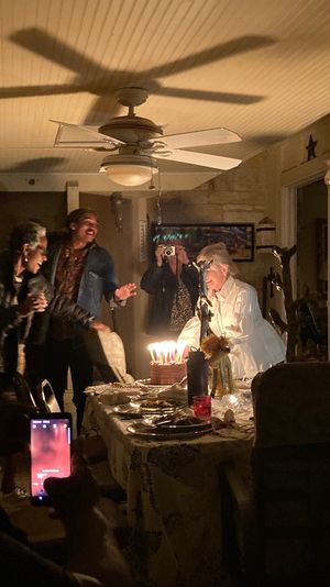 Cozy indoor birthday party at home — friends gathered around a dining table as a person leans toward a lit cake with many candles, guests cheer and snap photos with smartphones beneath a ceiling fan.