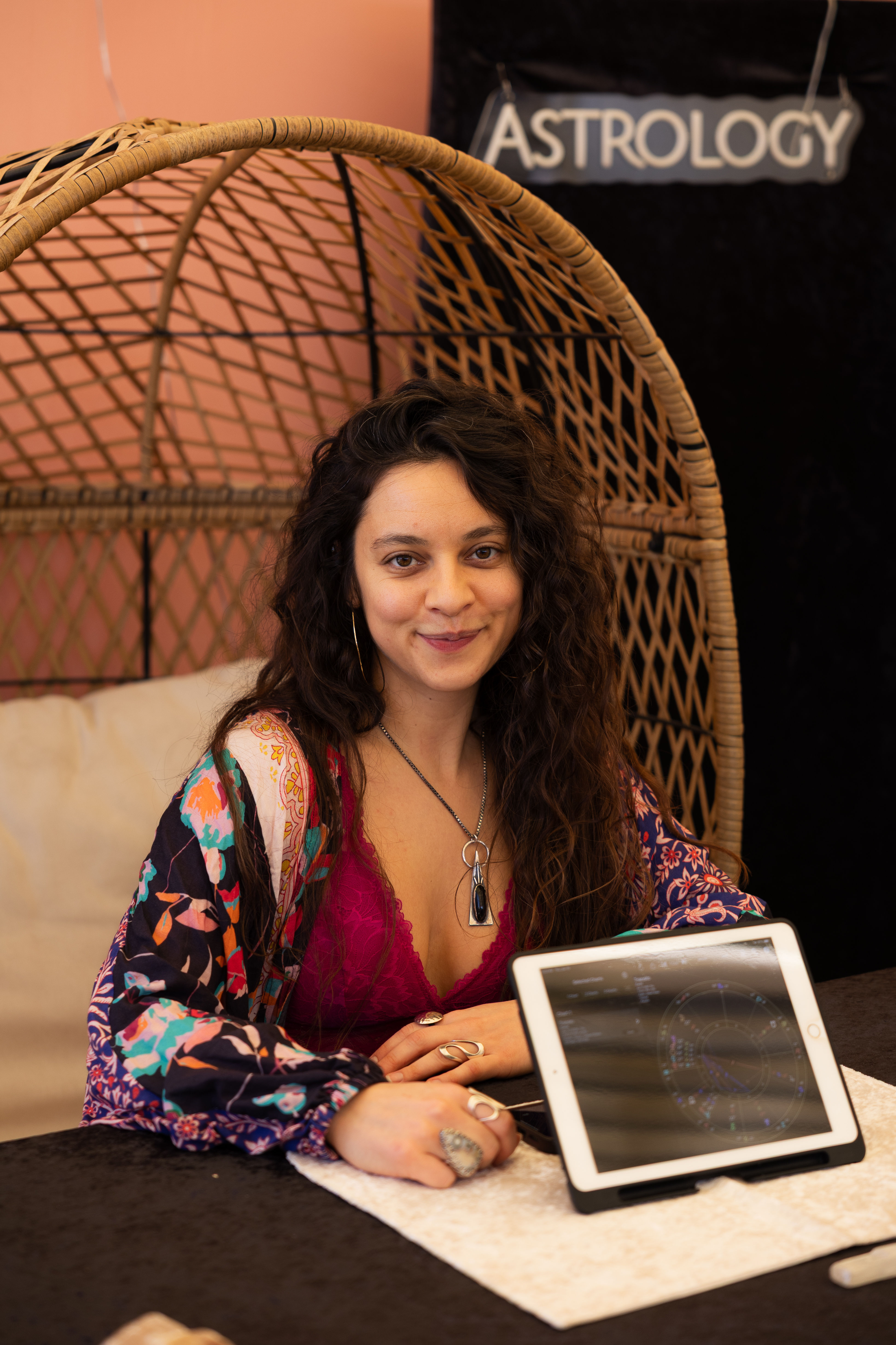 Smiling bohemian astrology reader seated in a wicker chair at a booth, wearing a colorful robe and pendant, with a tablet displaying a natal chart.