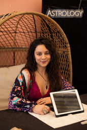 Smiling bohemian astrology reader seated in a wicker chair at a booth, wearing a colorful robe and pendant, with a tablet displaying a natal chart.