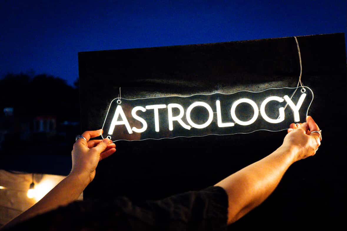 Hands holding a glowing white neon "ASTROLOGY" sign against a deep blue night sky