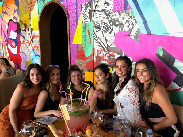 Six friends smiling at a restaurant table with a large fruit punch bowl, neon bendy straws and wine glasses, seated in front of a colorful graffiti mural wall.