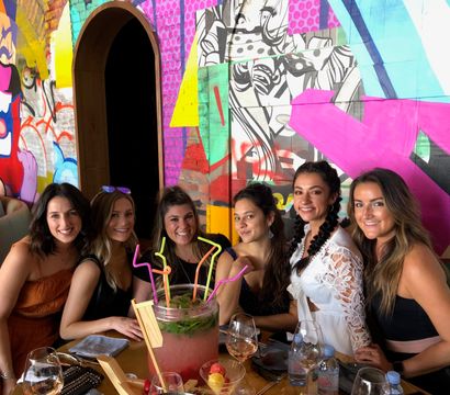 Six friends smiling at a restaurant table with a large fruit punch bowl, neon bendy straws and wine glasses, seated in front of a colorful graffiti mural wall.