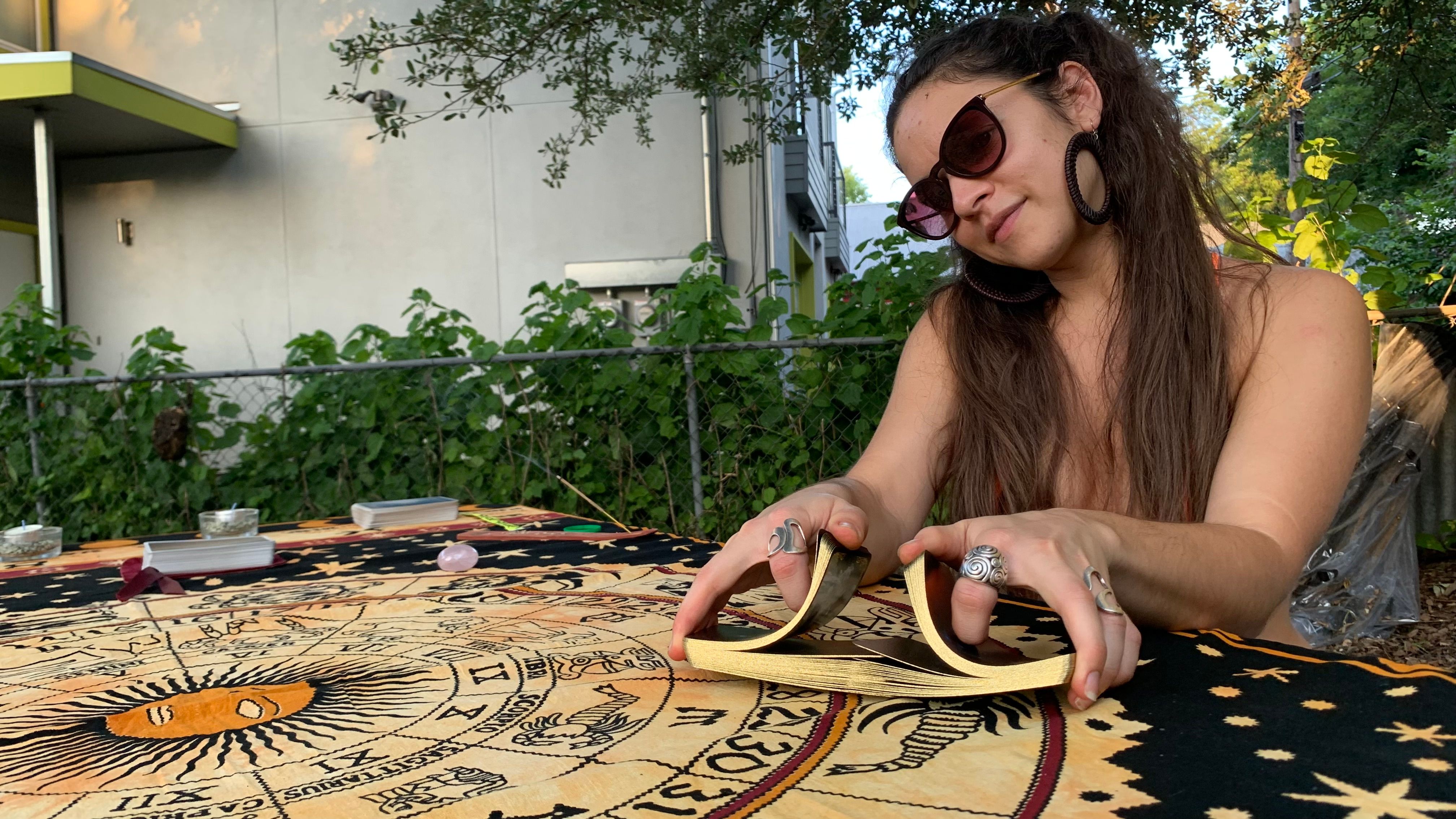 Person shuffling gold-edged tarot cards over an astrology tapestry with sun and zodiac symbols at an outdoor backyard table, wearing sunglasses, large hoop earrings and statement rings.