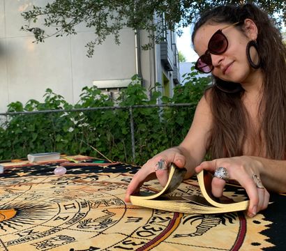 Person shuffling gold-edged tarot cards over an astrology tapestry with sun and zodiac symbols at an outdoor backyard table, wearing sunglasses, large hoop earrings and statement rings.
