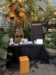 Astrology pop-up booth in a lush greenhouse: black-draped table with tablet displaying a birth chart, illuminated 'ASTROLOGY' sign, lightbox reading sign, string lights, wooden stool and surrounding potted plants.