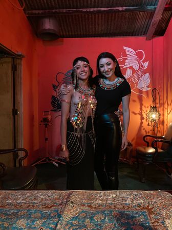 Two smiling women wearing embellished Egyptian-style beaded necklaces and a sequined costume, posing in a dim red-walled lounge with a koi mural, ornate lamp, vintage chairs and a patterned tablecloth in the foreground.