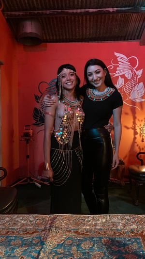 Two smiling women wearing embellished Egyptian-style beaded necklaces and a sequined costume, posing in a dim red-walled lounge with a koi mural, ornate lamp, vintage chairs and a patterned tablecloth in the foreground.