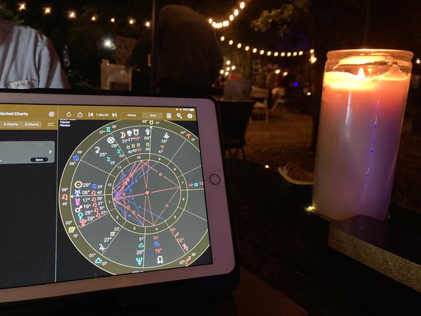 Tablet showing a colorful astrology chart on a candlelit outdoor patio table at night, warm string lights and a large white pillar candle in the background.