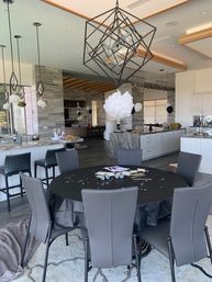 Modern open-plan kitchen and dining area set for a black-and-white party: round table with black tablecloth, gray chairs, confetti and party favors, geometric pendant light and white paper pom-pom, marble island and bar stools, black-and-white balloons.