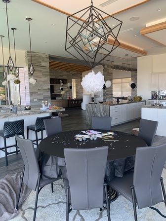Modern open-plan kitchen and dining area set for a black-and-white party: round table with black tablecloth, gray chairs, confetti and party favors, geometric pendant light and white paper pom-pom, marble island and bar stools, black-and-white balloons.