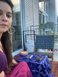 Person on a city apartment balcony with a boho setup: blue-patterned tablecloth holding a lit candle, a tarot/oracle deck and wooden box, woven chair, potted trailing plant and sliding glass door reflecting nearby buildings.