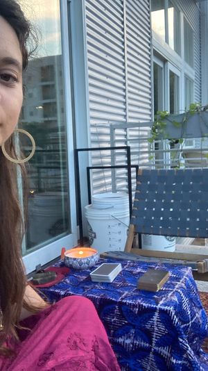 Person on a city apartment balcony with a boho setup: blue-patterned tablecloth holding a lit candle, a tarot/oracle deck and wooden box, woven chair, potted trailing plant and sliding glass door reflecting nearby buildings.