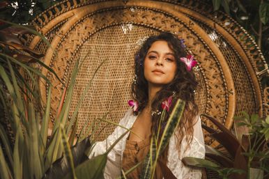 Boho-chic portrait of a woman with flowers in her hair seated in a woven peacock chair surrounded by lush tropical plants and pink orchids