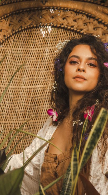 Boho-chic portrait of a woman with flowers in her hair seated in a woven peacock chair surrounded by lush tropical plants and pink orchids
