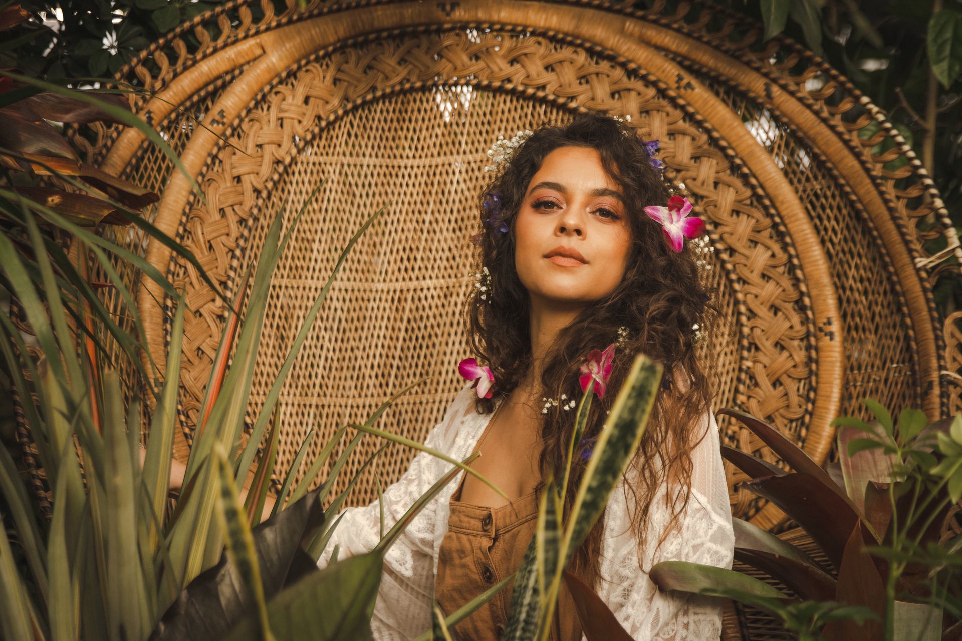 Boho-chic portrait of a woman with flowers in her hair seated in a woven peacock chair surrounded by lush tropical plants and pink orchids
