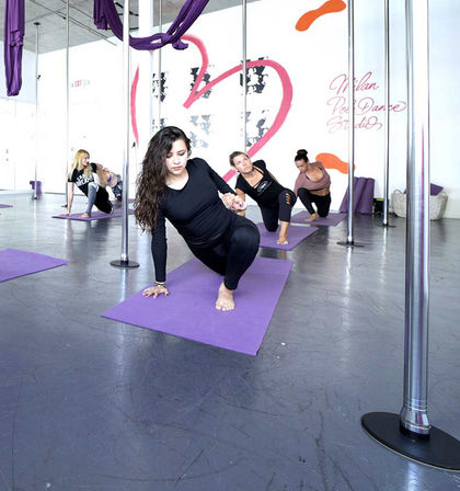 Focused women practicing floor stretches in a bright pole-fitness studio on purple mats with poles and aerial silks visible — energetic group class