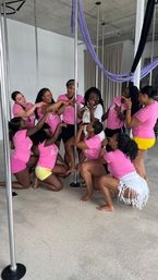 Group of women in pink shirts posing around a pole in an indoor pole-fitness studio with purple aerial silks, celebrating a surprised woman in white.