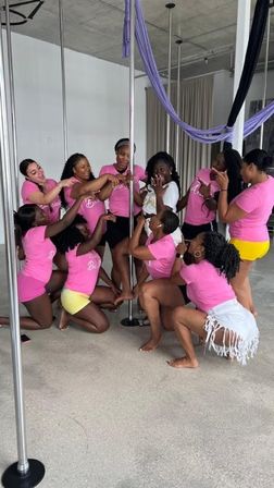 Group of women in pink shirts posing around a pole in an indoor pole-fitness studio with purple aerial silks, celebrating a surprised woman in white.