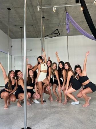 Smiling group of women in activewear posing around a pole inside a bright pole-fitness and aerial yoga studio with hanging silks and exposed concrete ceiling