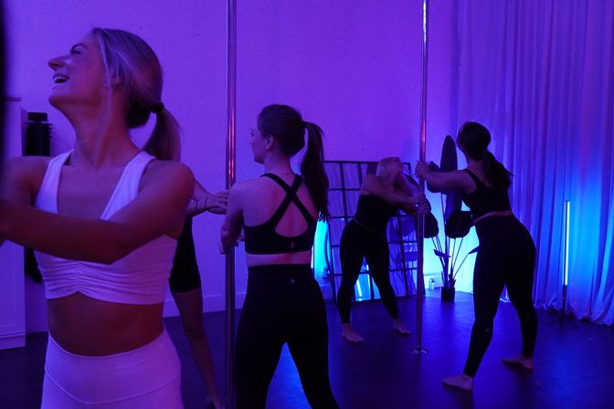 Group of women in a pole fitness class practicing moves and stretching around vertical poles in a purple-blue lit indoor studio