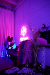 Neon-lit indoor lounge corner bathed in purple-pink light with a glowing wall neon sign, feather boas, faux cash, decorative grasses and a couch.