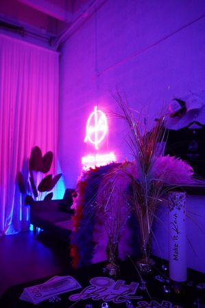 Neon-lit indoor lounge corner bathed in purple-pink light with a glowing wall neon sign, feather boas, faux cash, decorative grasses and a couch.
