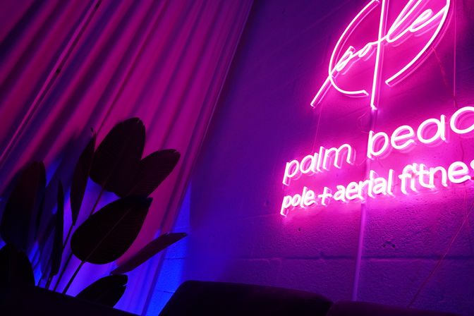 Vibrant magenta neon sign advertising pole and aerial fitness glows on a textured wall, with draped curtains and silhouetted tropical plant leaves bathed in pink and blue ambient light.