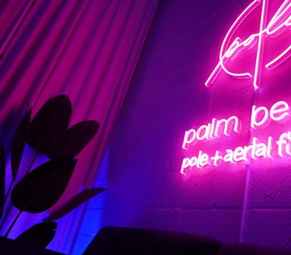 Vibrant magenta neon sign advertising pole and aerial fitness glows on a textured wall, with draped curtains and silhouetted tropical plant leaves bathed in pink and blue ambient light.