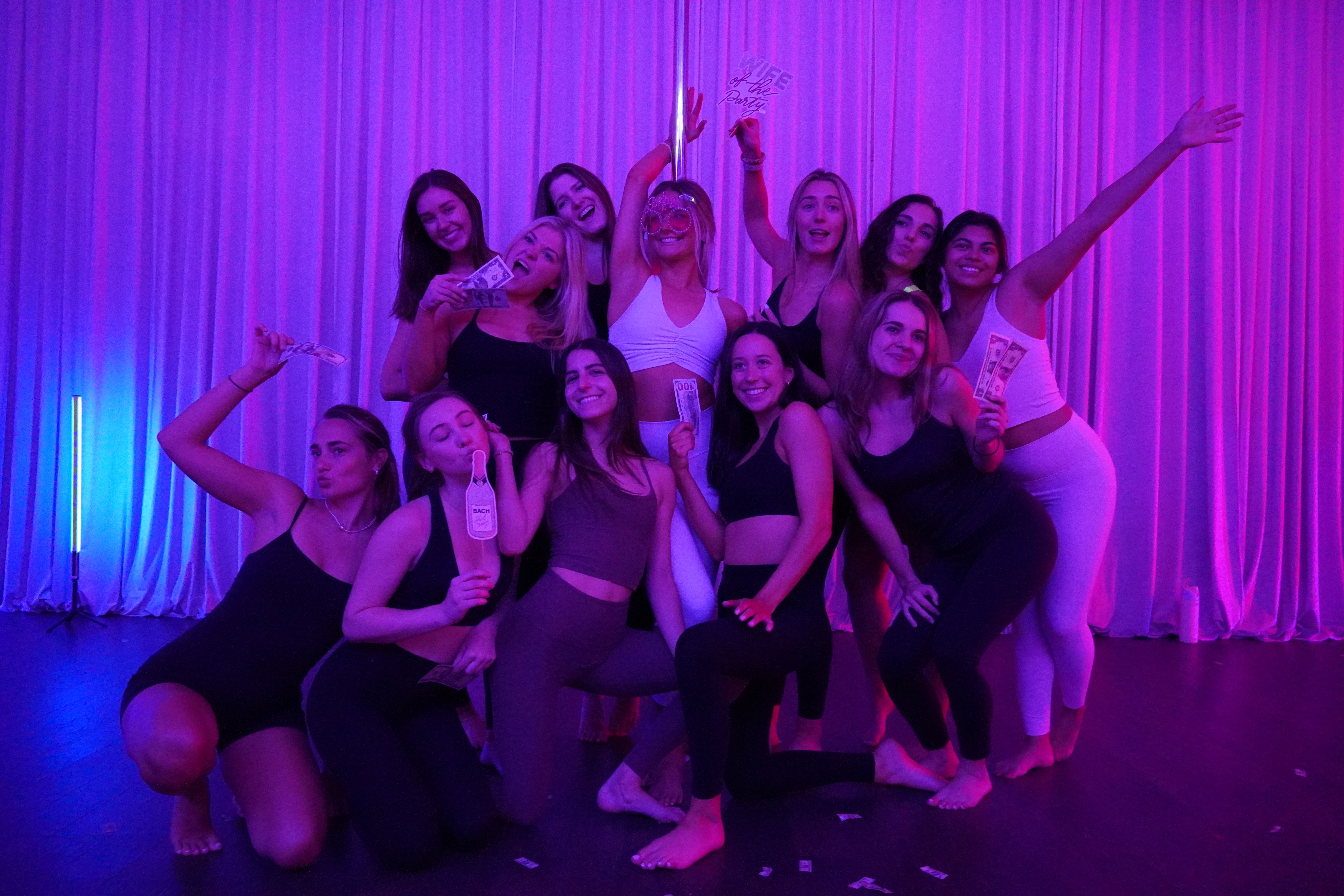 Group of a dozen women in activewear posing and smiling at a neon purple-lit dance studio bachelorette party, holding play money and a novelty bottle in front of floor-to-ceiling draped curtains.