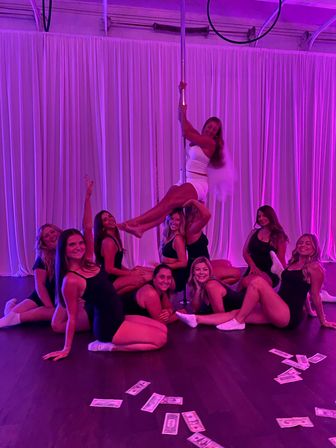 Group of women in athletic wear posing around a pole in a pink-purple lit dance studio for a bachelorette pole-fitness celebration, dollar bills scattered on the hardwood floor.